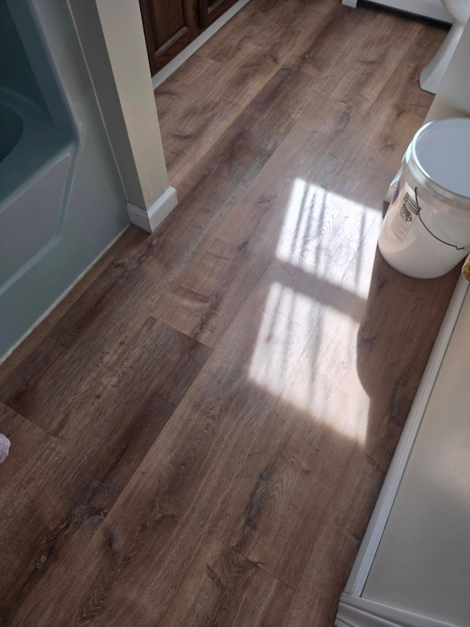 Hallway with wood-look flooring, sunlight streaming across it, and a white paint bucket near the wall
