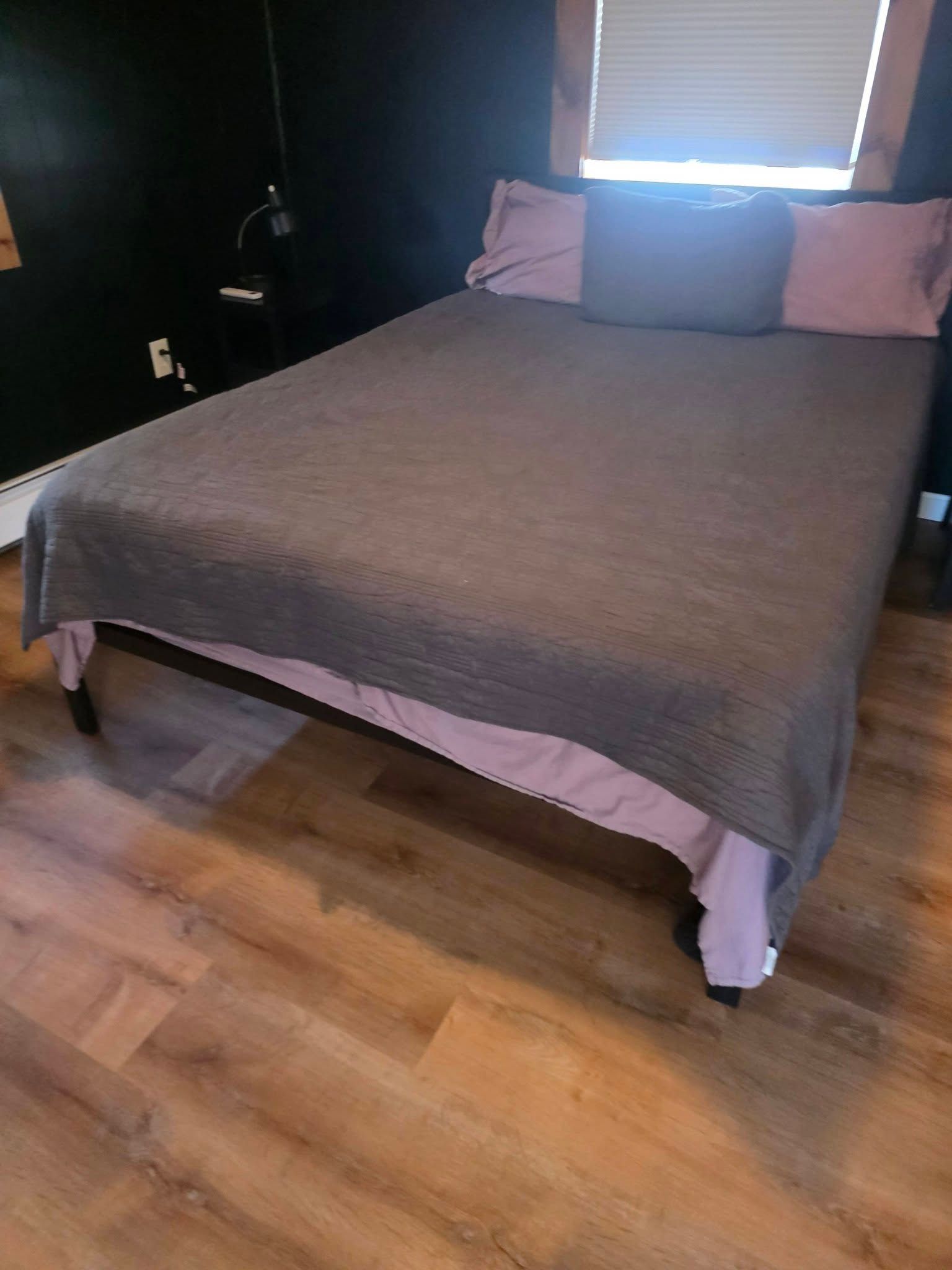 Gray bed with pink pillows and blanket in a bedroom with wood flooring and a window blinds behind it