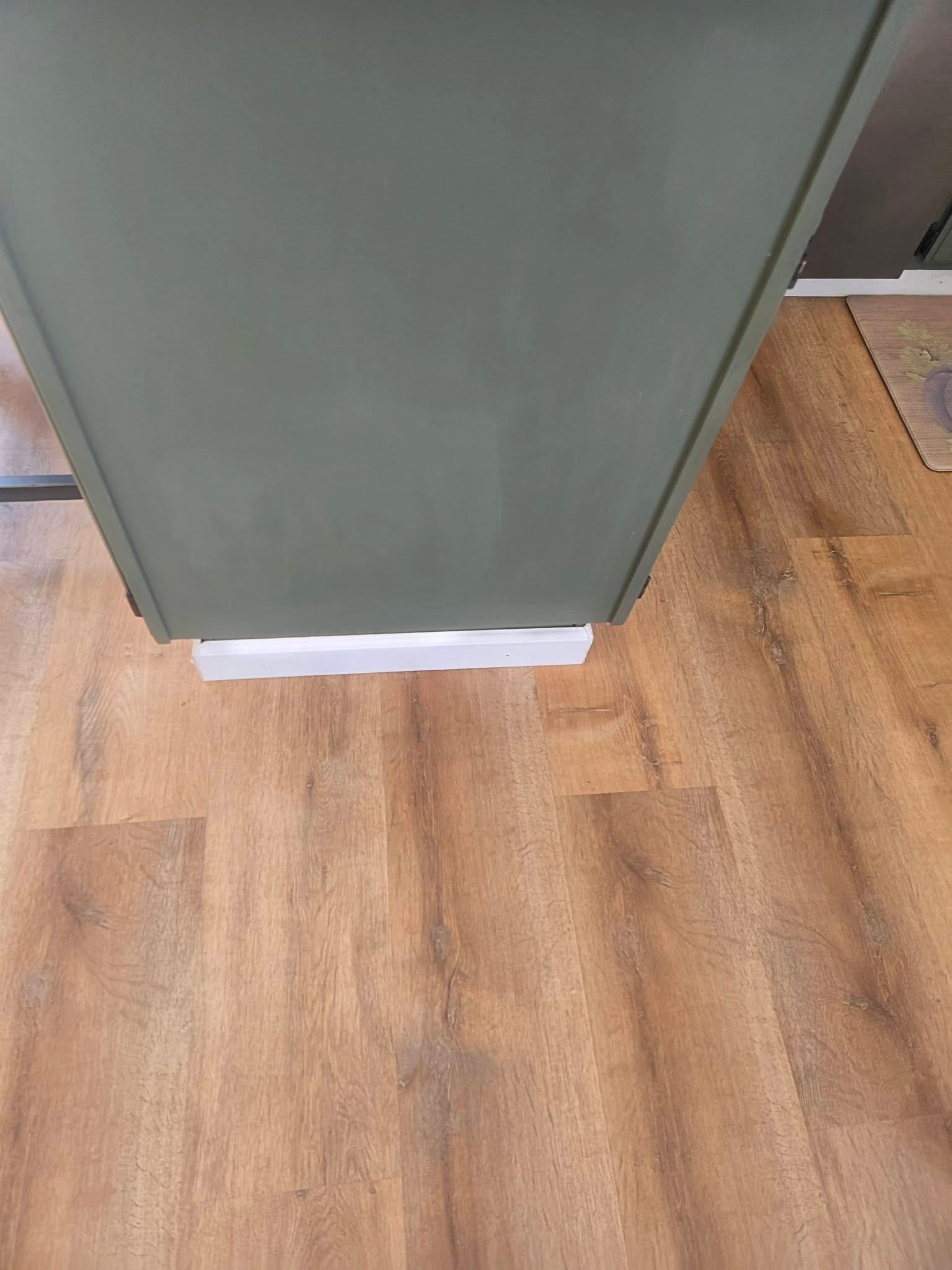 Green cabinet in a kitchen, with white base trim and wood-look flooring.