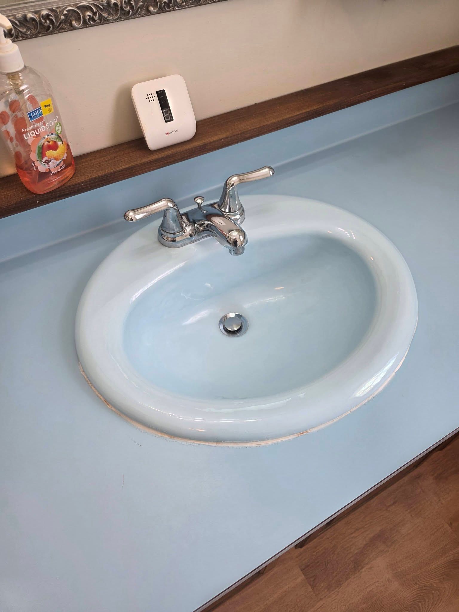 White oval bathroom sink with chrome faucet on a pale blue countertop and wooden floor trim.
