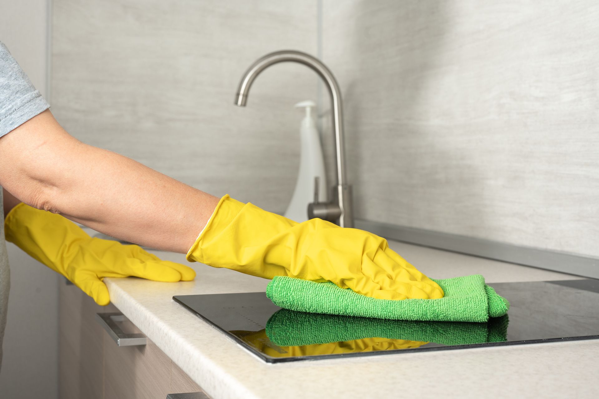 Gloved hands wiping a kitchen countertop beside a sink with a green sponge