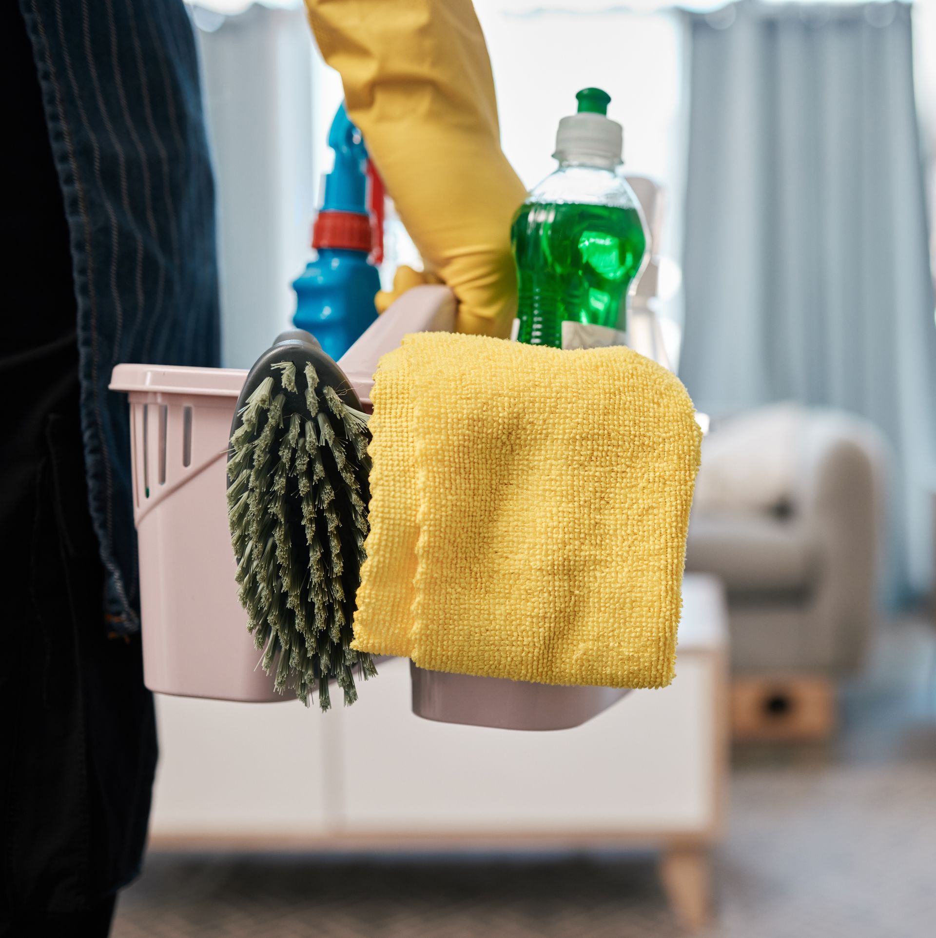 Cleaning caddy with spray bottles, sponge, and scrub brush held by a gloved hand in a room