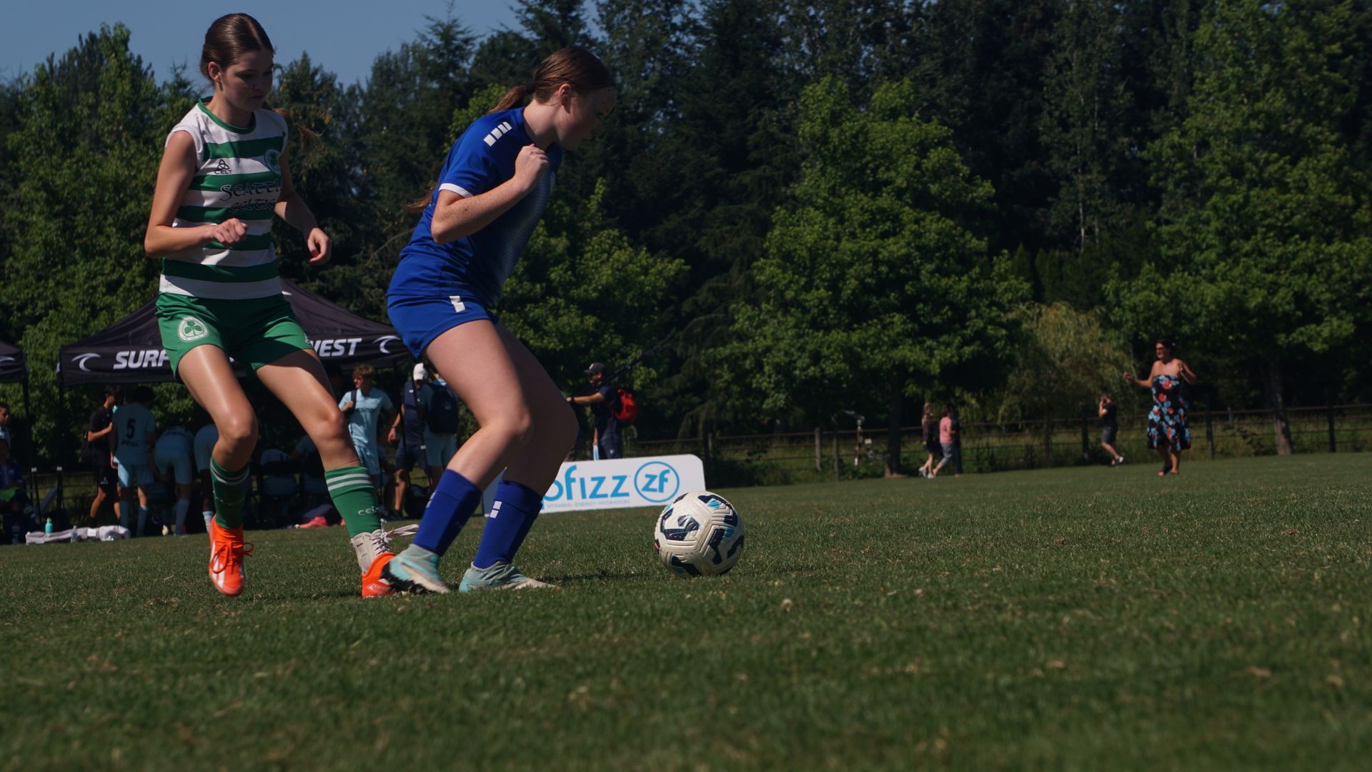 Two girls are playing soccer on a field with trees in the background.