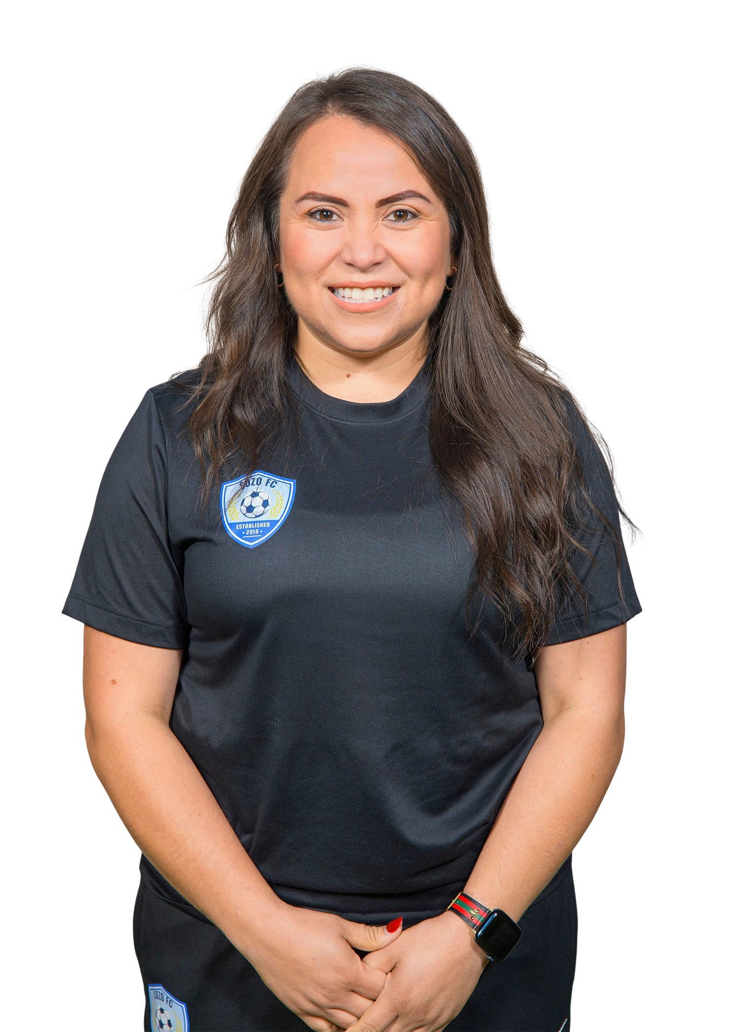 Woman in a black soccer shirt smiling. Hands clasped, long brown hair, and a smartwatch.