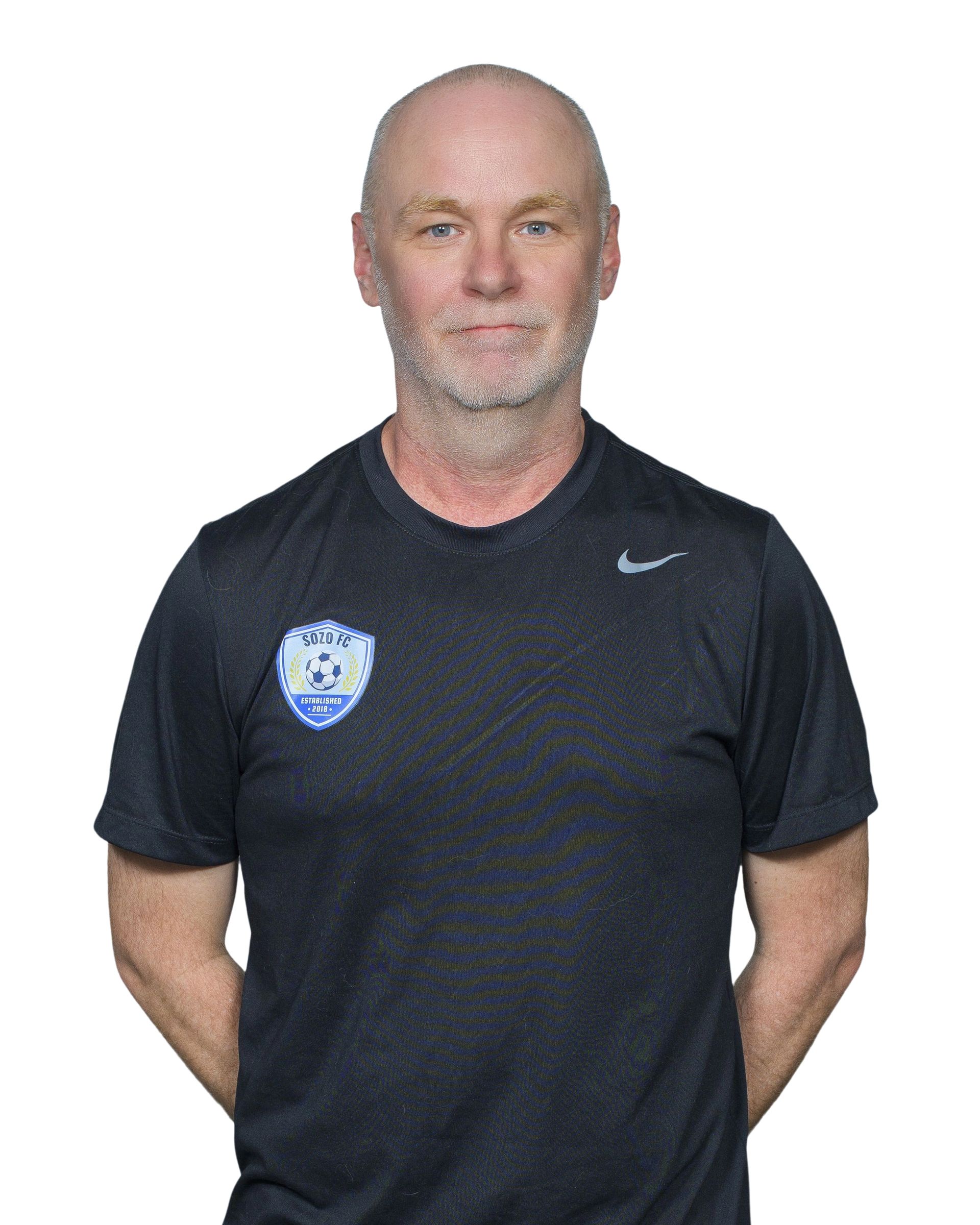 A man with a beard is wearing a black polo shirt and smiling.