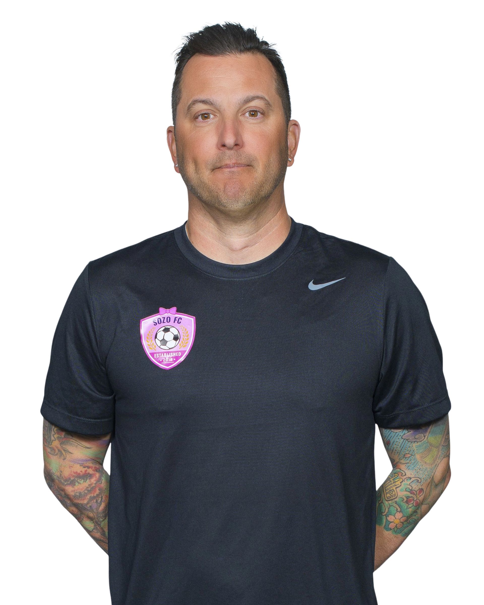 A man with a beard and tattoos is wearing a white shirt.