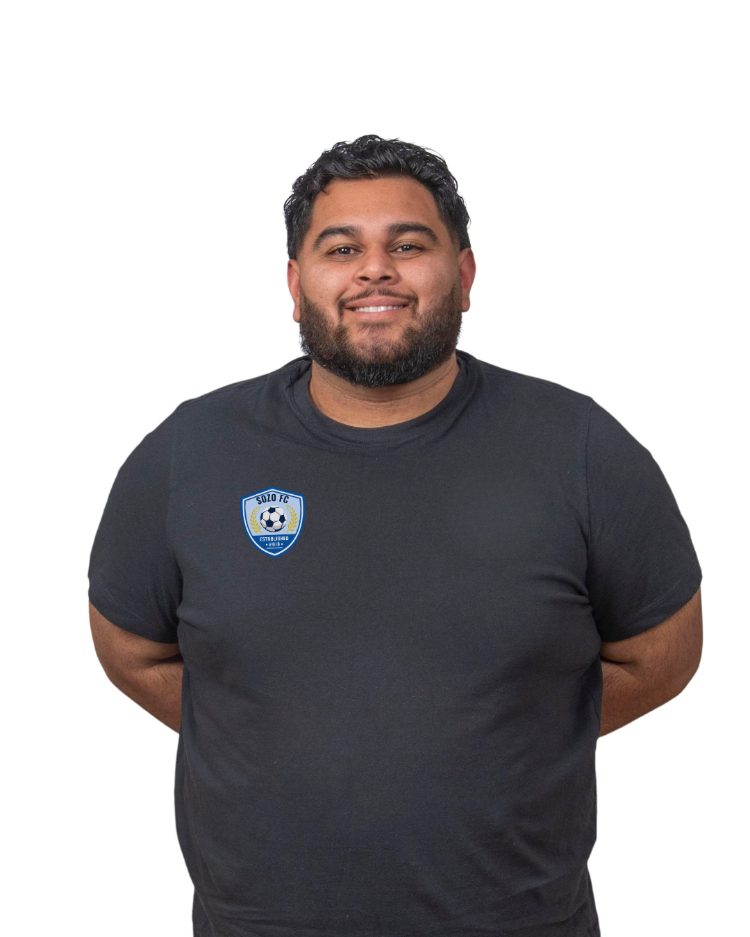 Person in a dark shirt standing against a white background, smiling with hands behind their back.