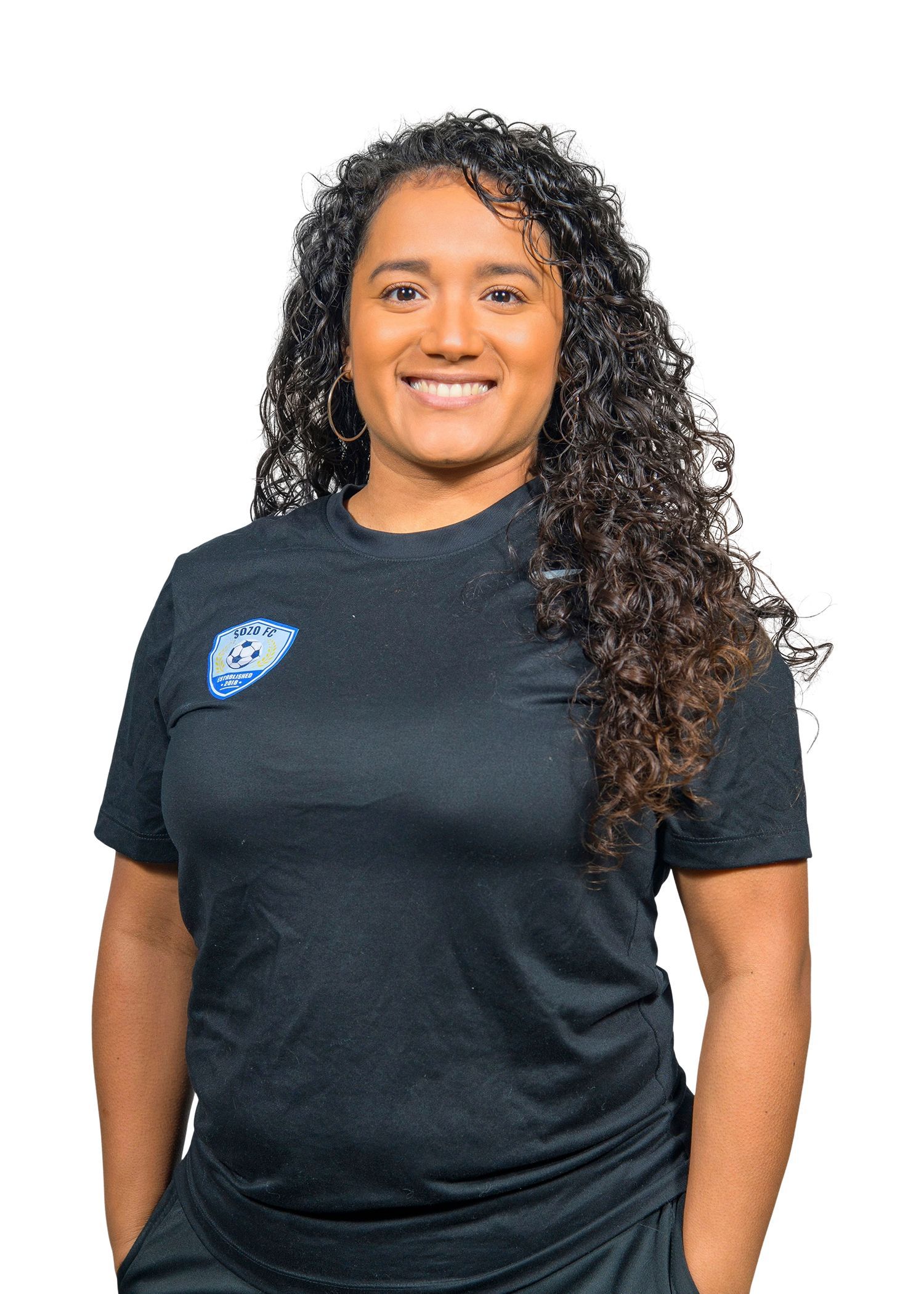 Woman with curly hair in a black shirt smiling, hands in pockets.