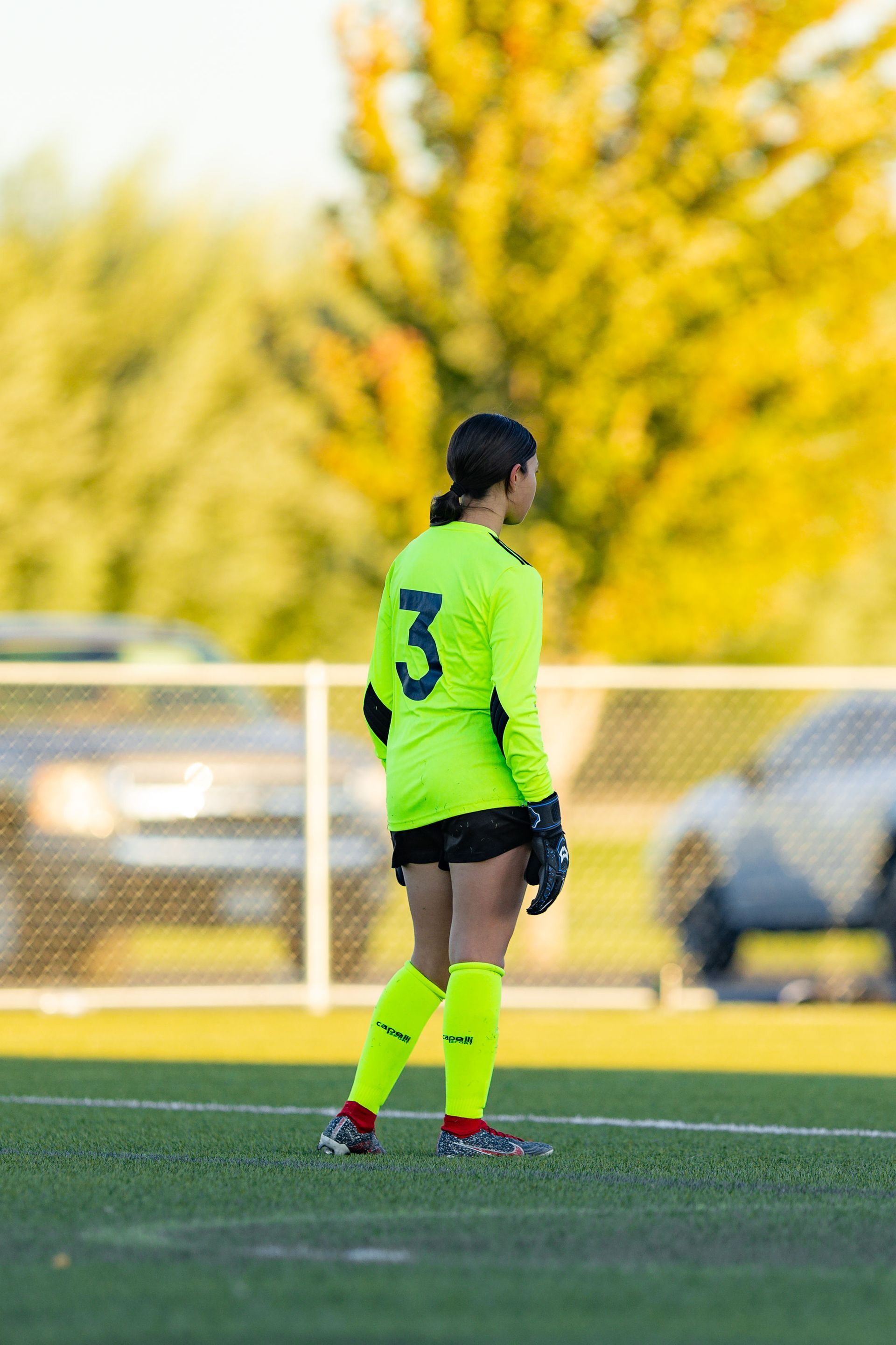 A female soccer player wearing a neon green jersey with the number 3 on it is standing on a field.