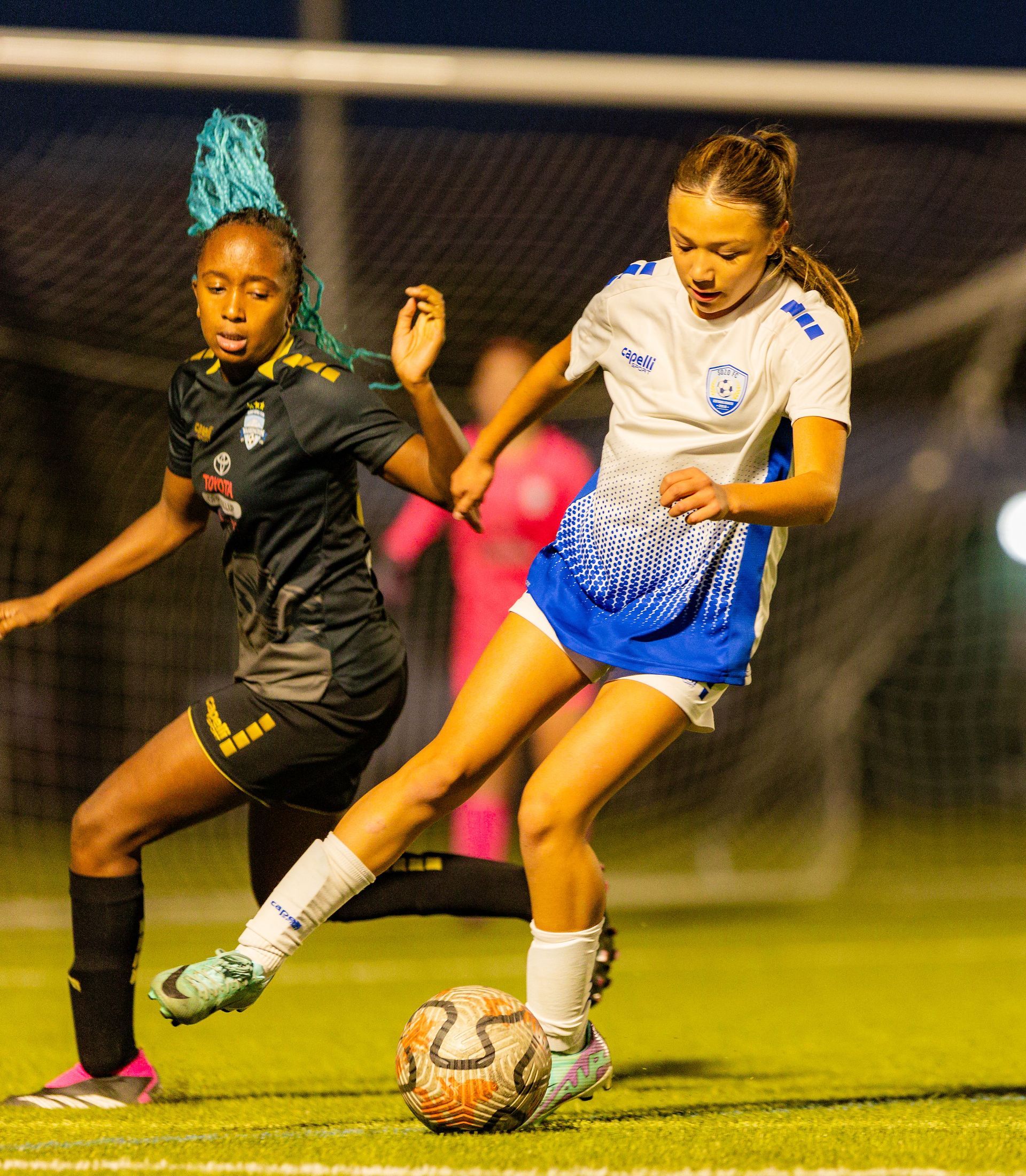 Two girls are playing soccer on a field and one has blue hair