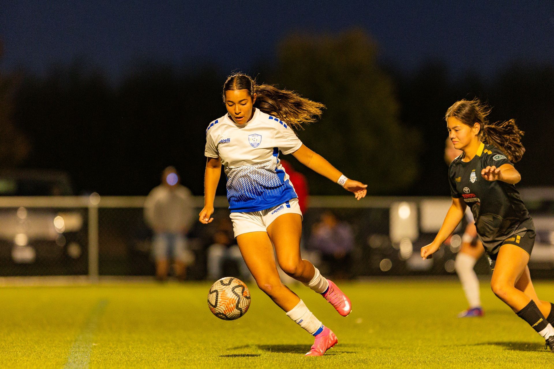 Two girls are playing soccer on a field at night.