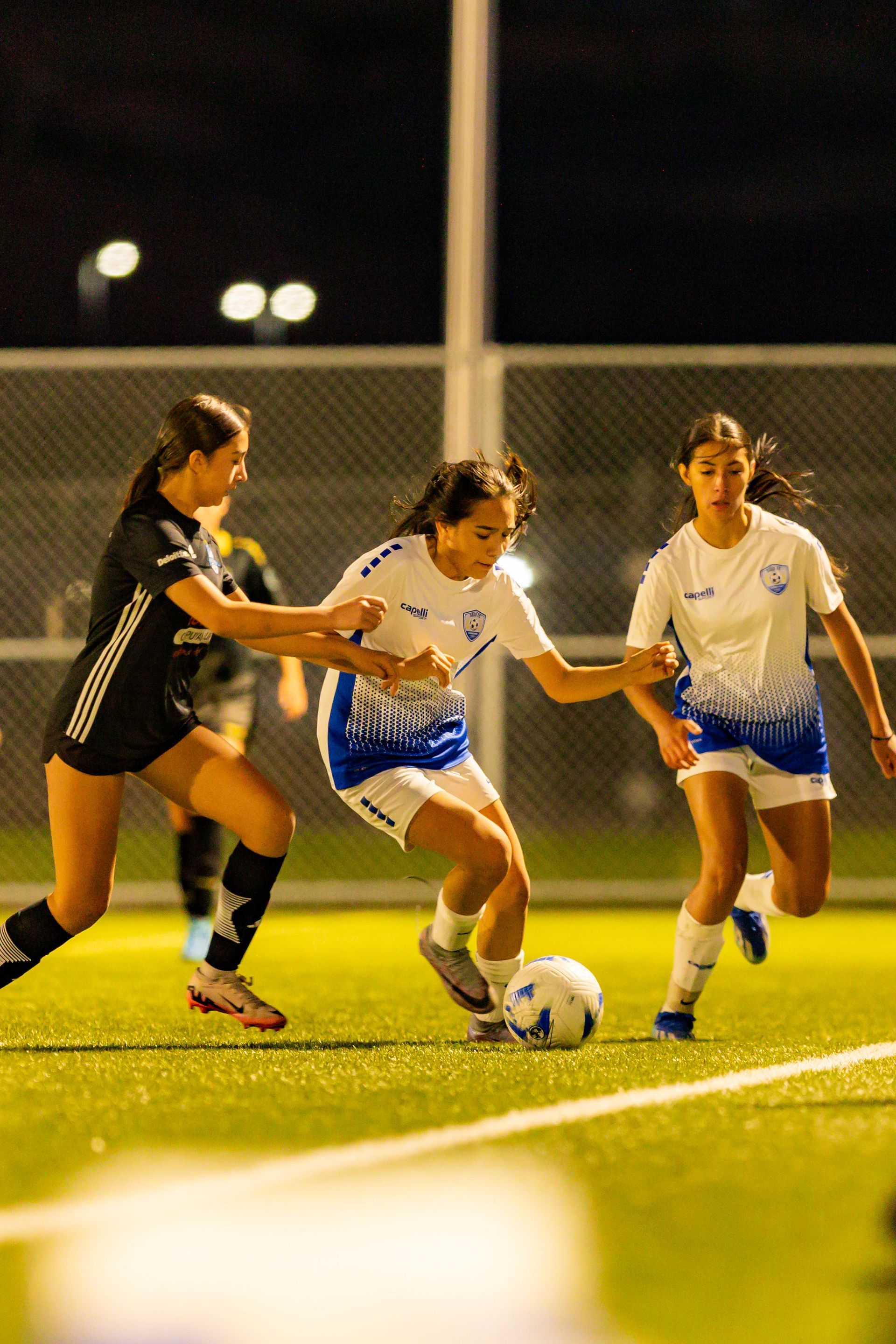 Three women are playing soccer on a field at night.
