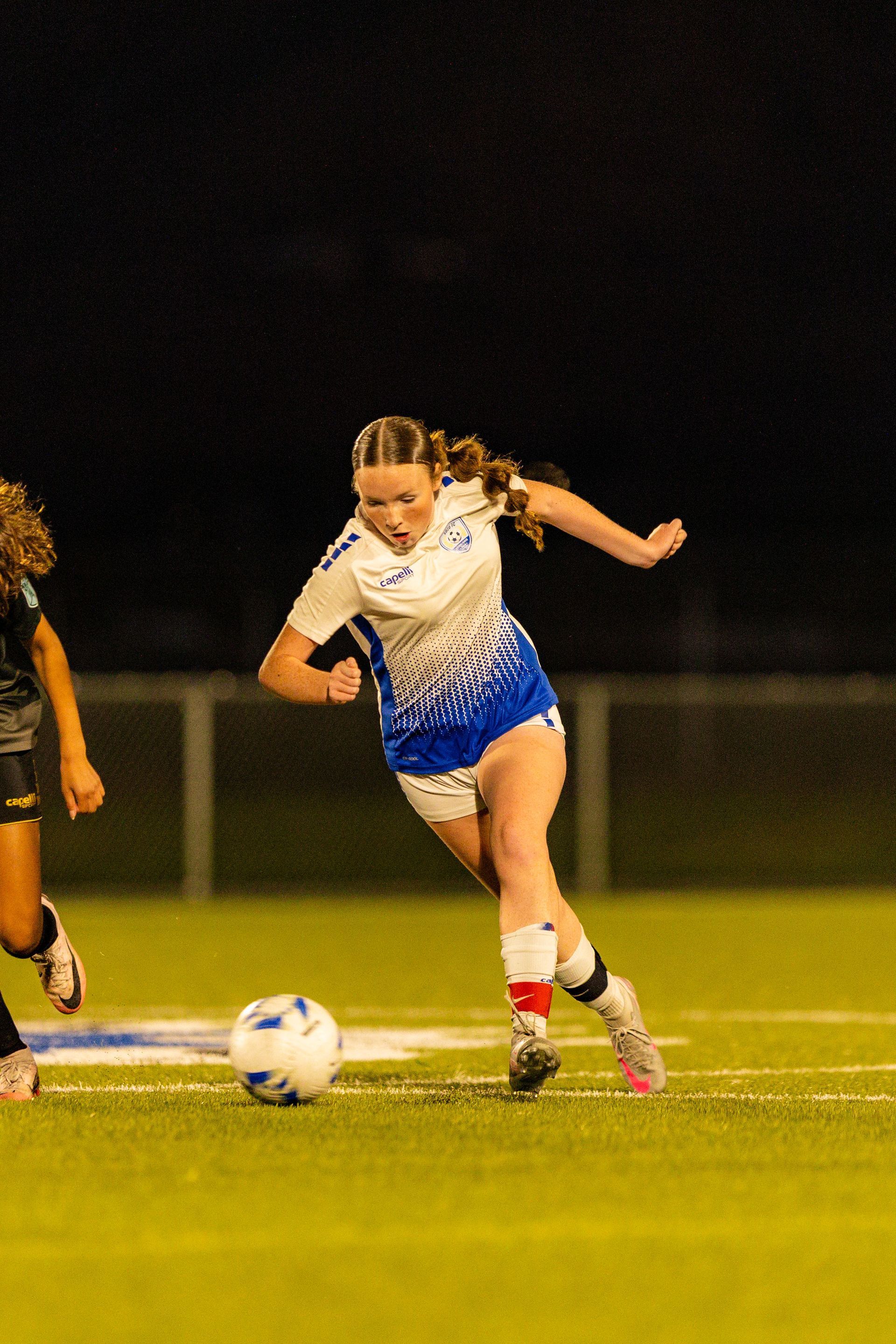 Two girls are playing soccer on a field at night.