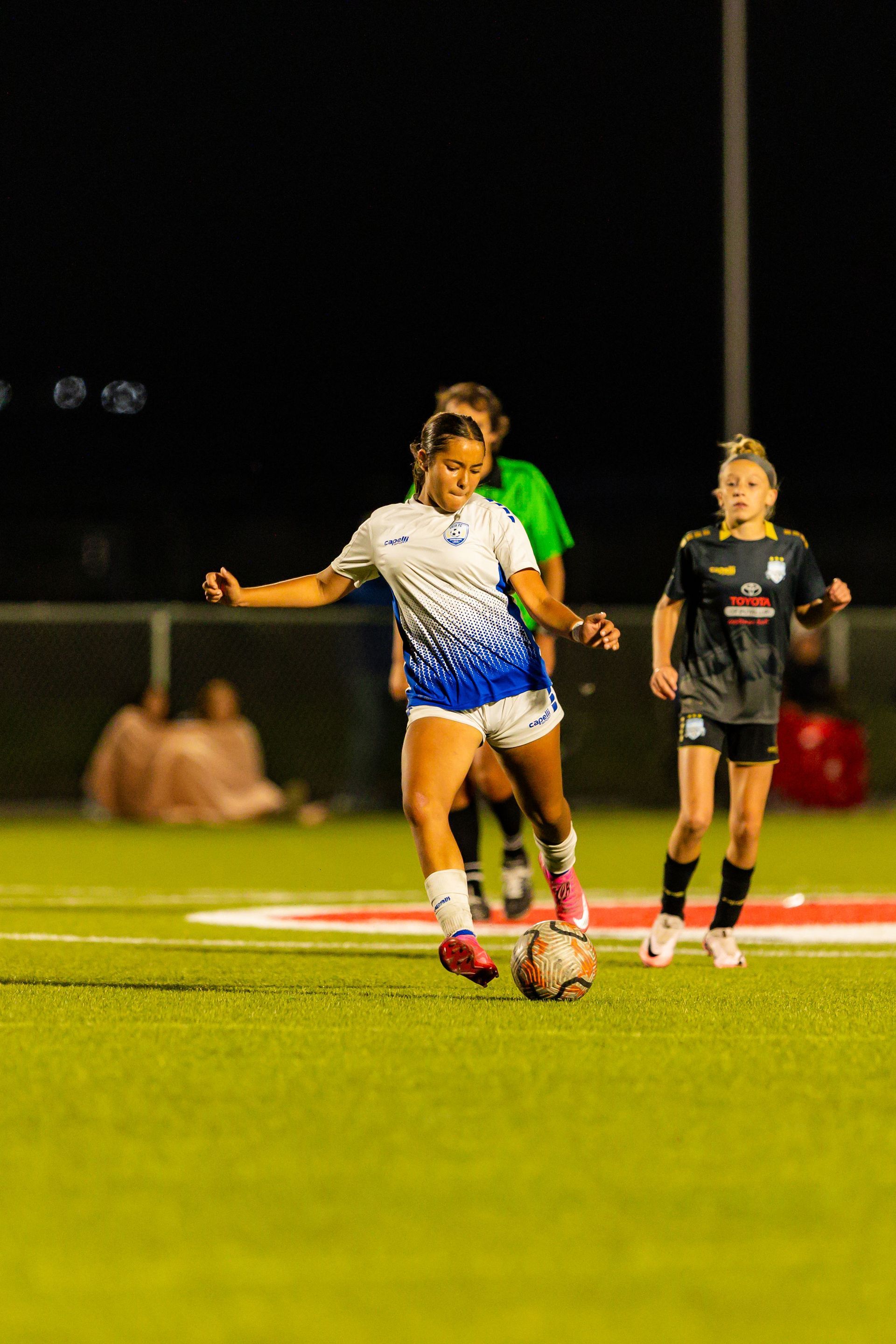 A woman is kicking a soccer ball on a field at night.