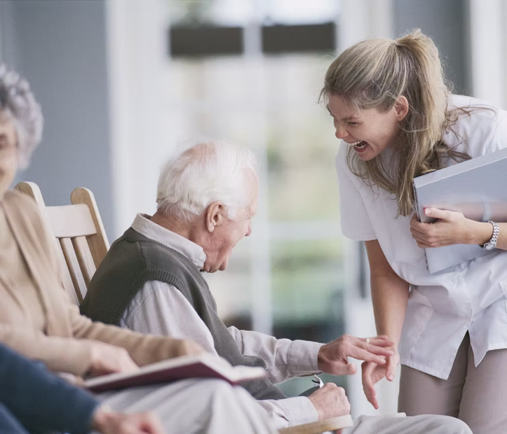 Caregiver smiling and leaning toward an elderly man in a bright nursing home lounge