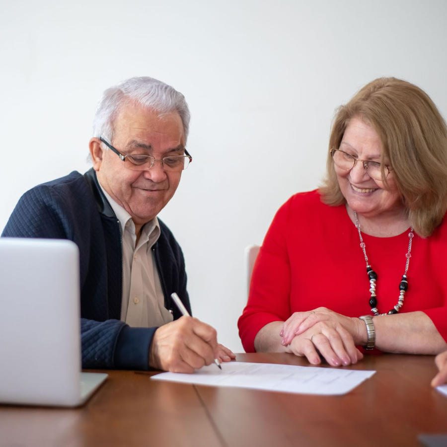 Two people reviewing documents at a desk beside a laptop, one writing and both smiling