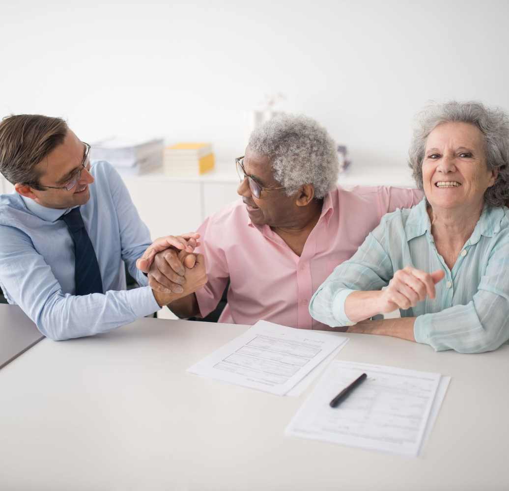 Three people at a table smiling and shaking hands over paperwork in a bright office