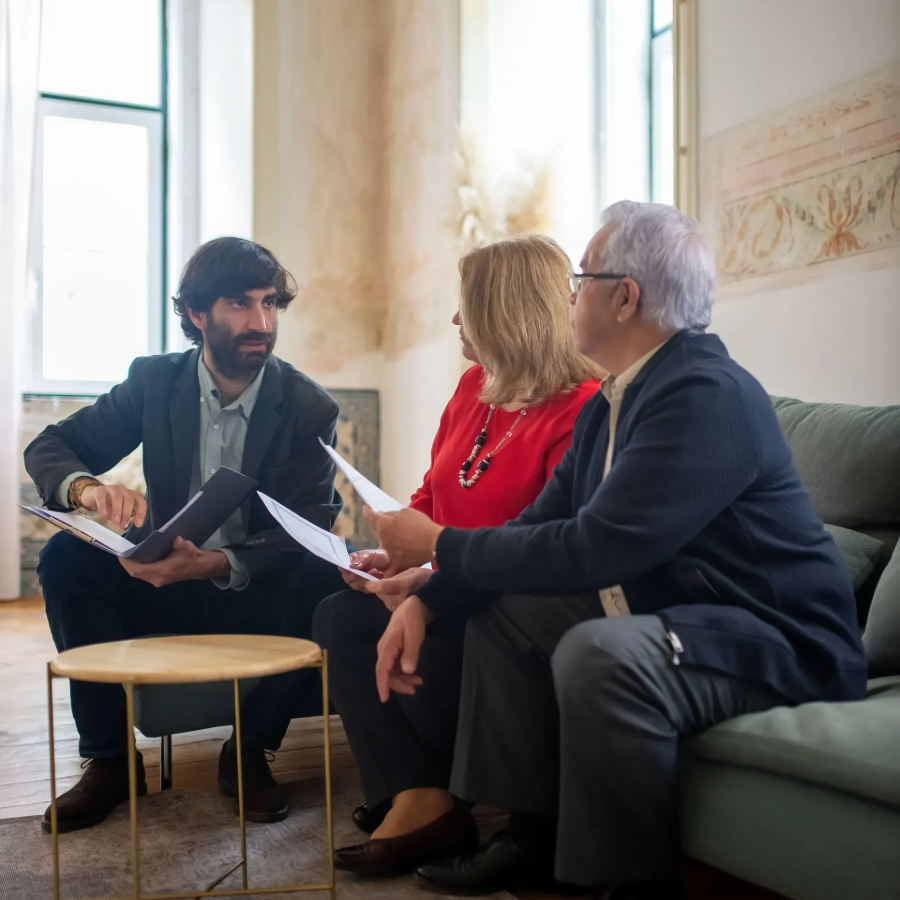 Three people in conversation on a sofa in a bright room, one holding papers.