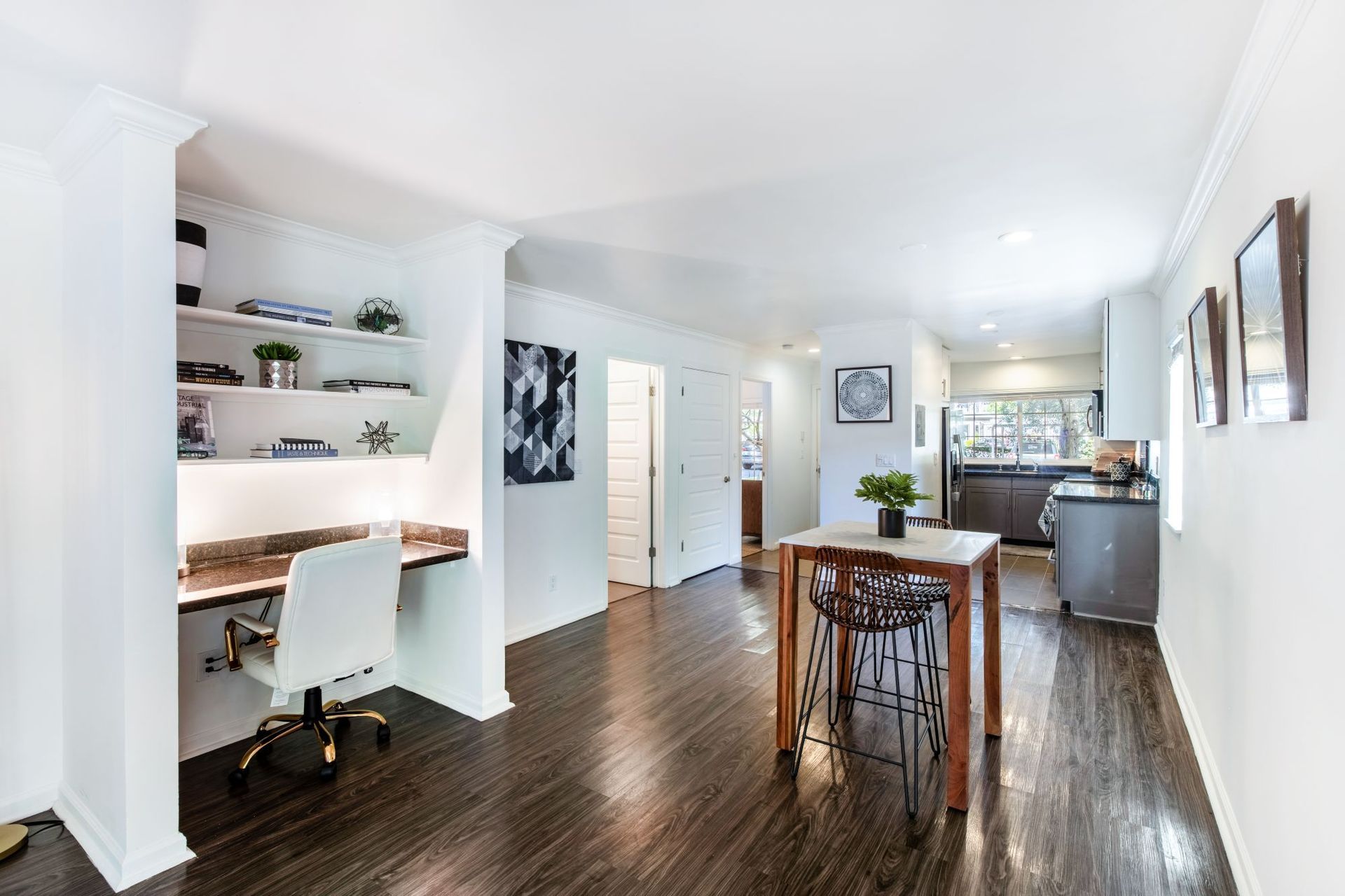 Bright open-concept living area with dark wood floors, white walls, desk nook, and kitchen in the background
