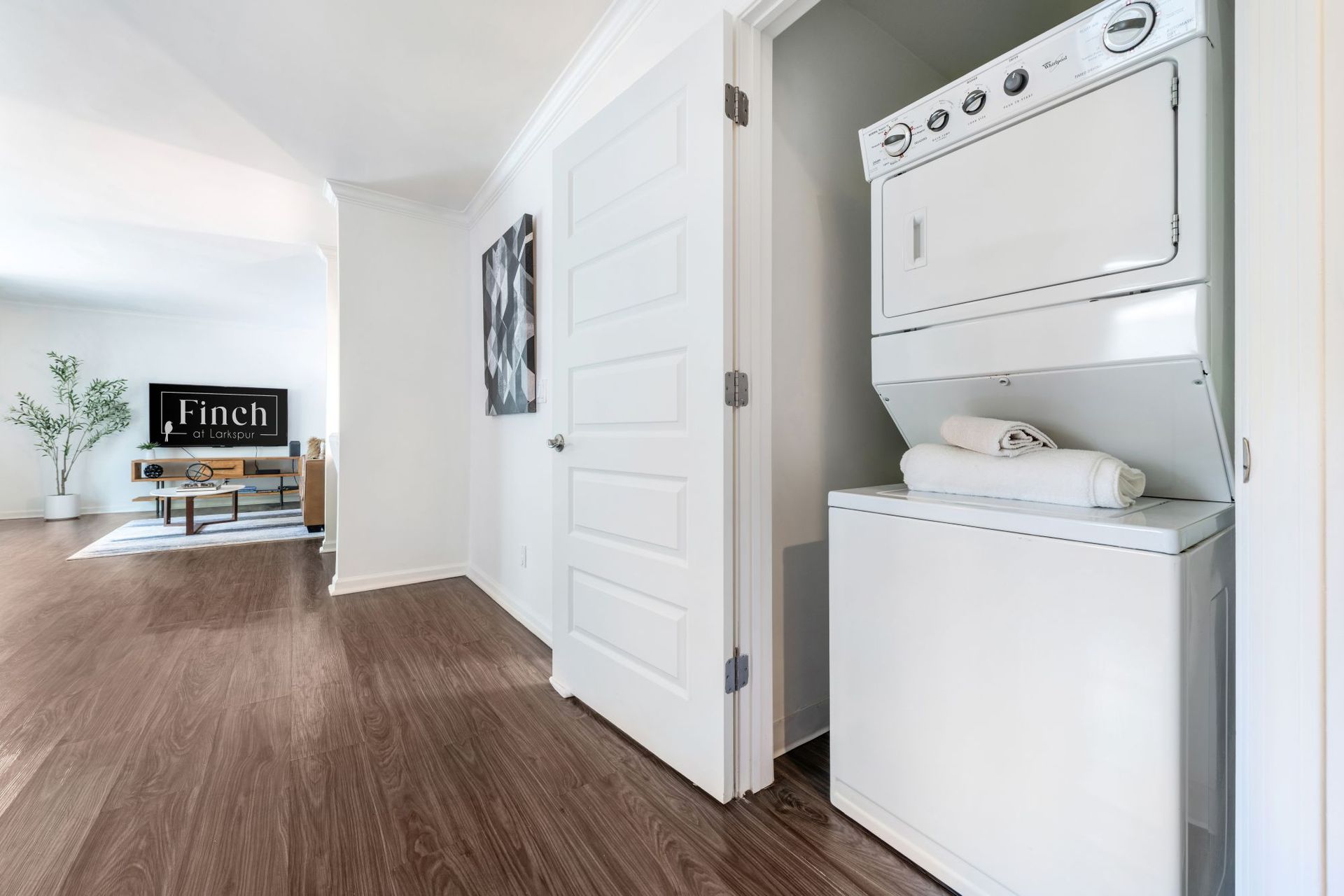 Hallway with stacked washer and dryer beside a white door, leading to a living room with wood floors.