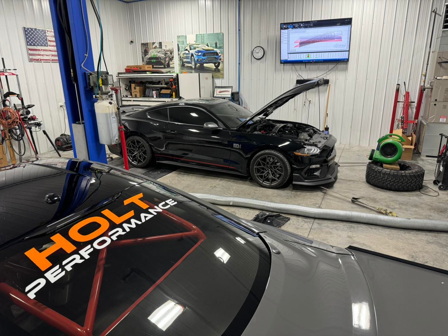 Two cars in a garage. One black, hood open; one grey with "HOLT PERFORMANCE" on windshield.