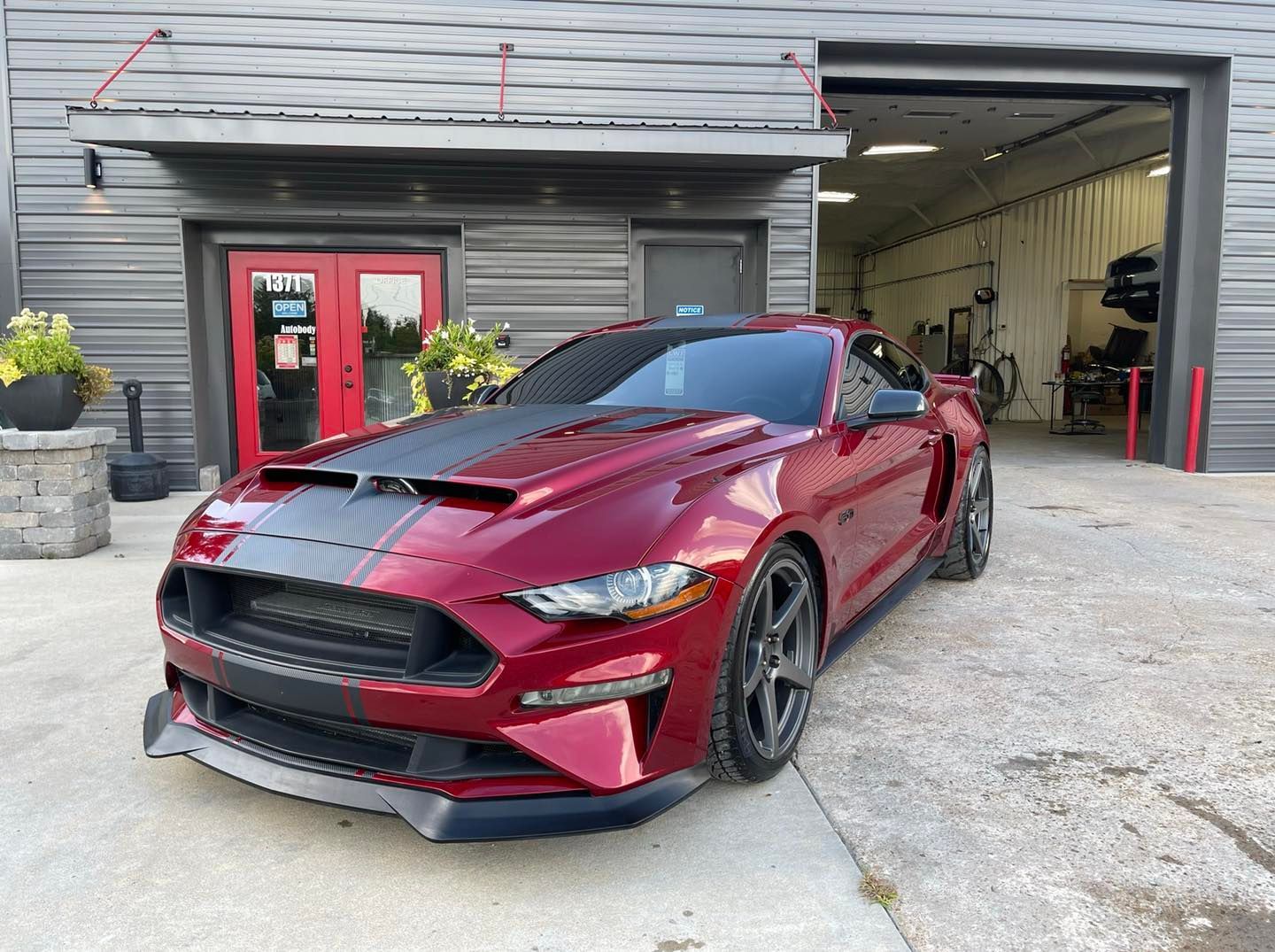 Red Mustang sports car parked outside a garage with a black hood stripe.