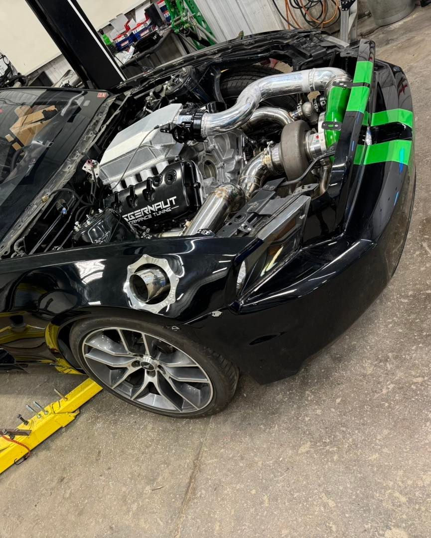 Black sports car with exposed engine, being worked on in a garage. Green tape highlights panels.