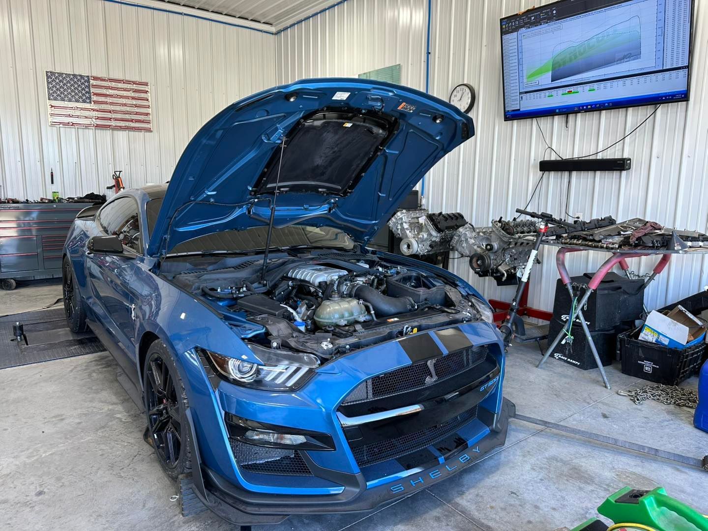 Blue Ford Mustang Shelby GT500 with hood open in a garage, U.S. flag on wall, dyno screen visible.
