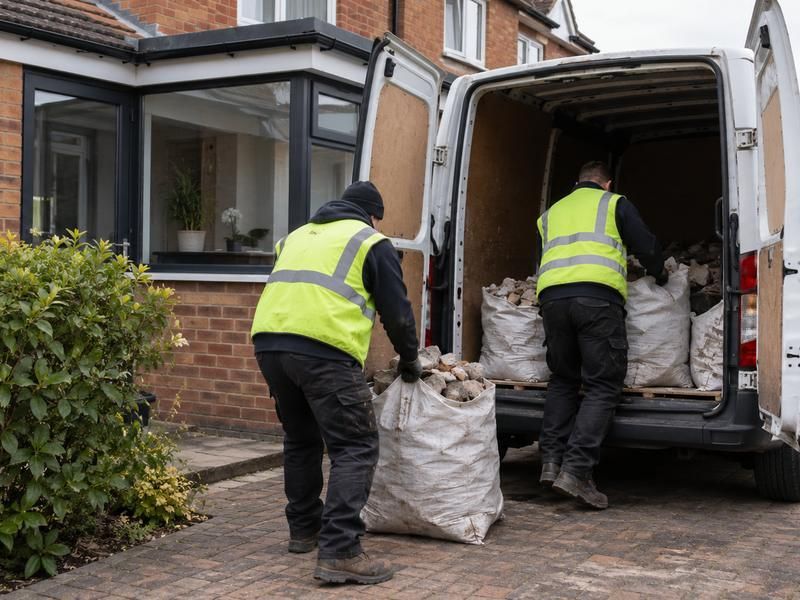 Two workers loading bags of rubble into a van outside a house