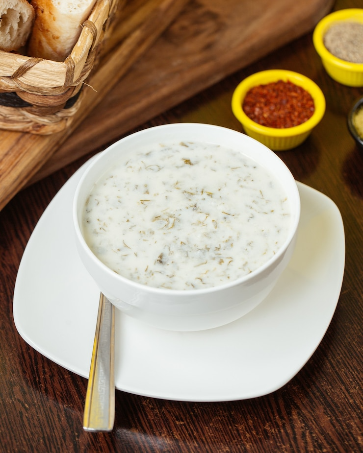 Bowl of creamy soup on a white saucer with spoon, on a wooden table with spices nearby