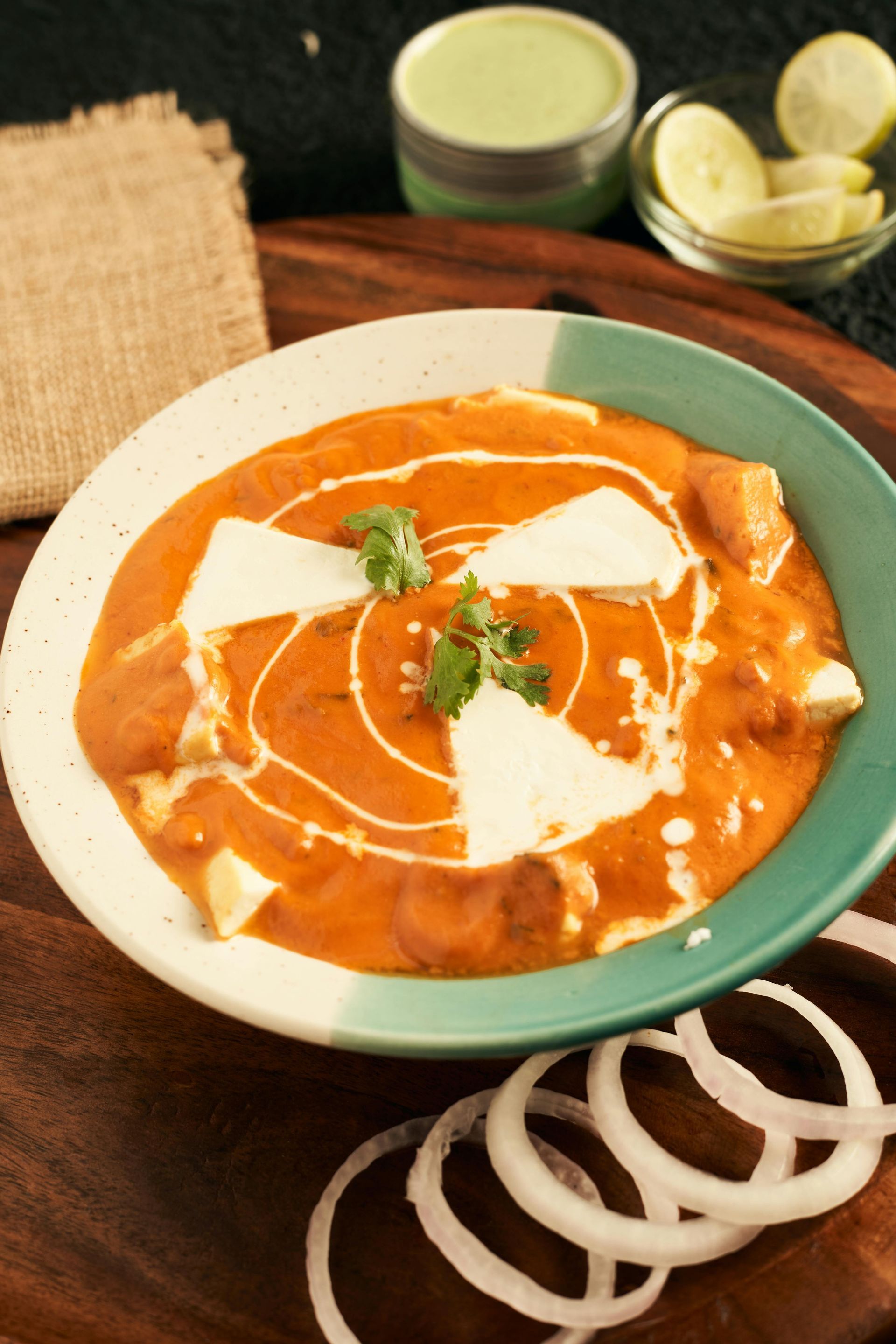 Paneer butter masala in a bowl with cream swirl, onion rings, and lime wedges on a table