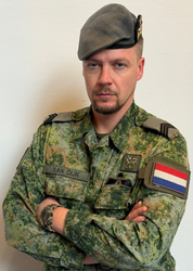 Gecamoufleerde soldaat die op een rots knielt en een geweer vasthoudt in een grasveld.