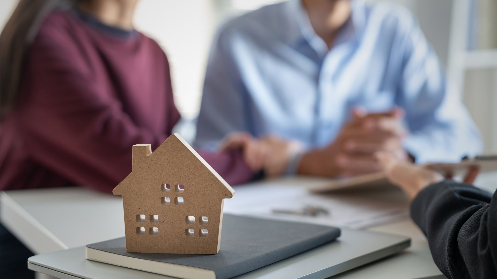 Wooden house model on a table with a couple and person, possibly discussing a property deal.