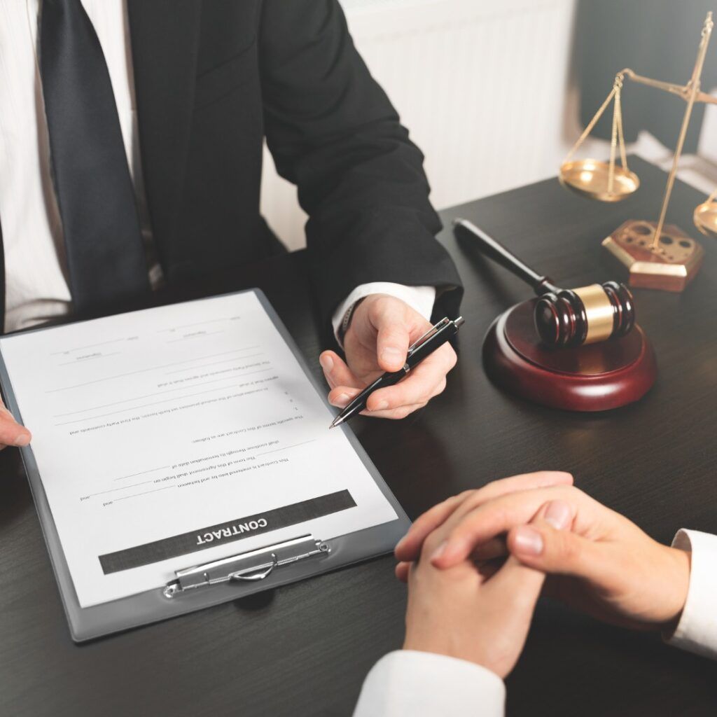 Lawyer pointing at contract for client to sign, with gavel and scales of justice.