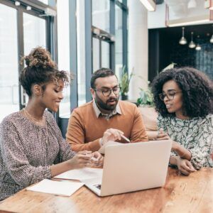 Three people collaborate at a table with a laptop. They point at the screen, discussing.