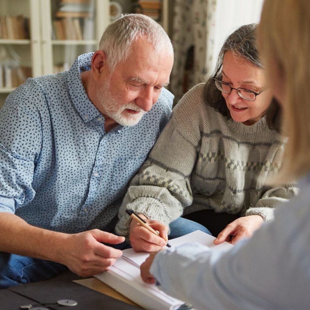 Older couple reviews paperwork, man signs document at table with another person.
