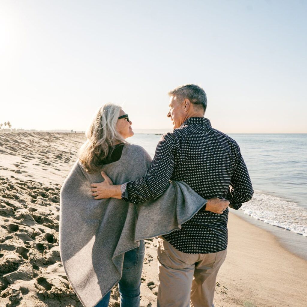 Couple walking on beach, arms around each other, looking at each other, sunny day.