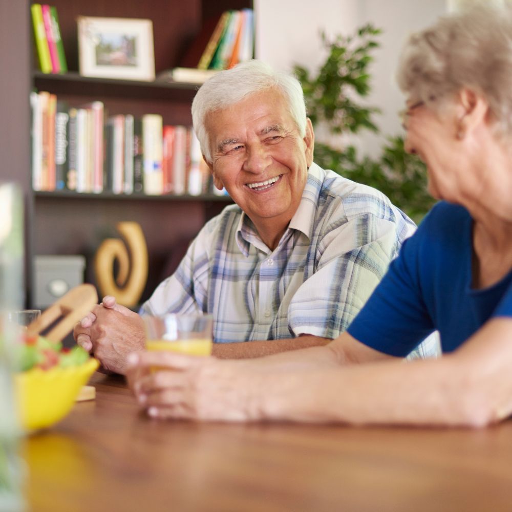 Smiling older person at a table, looking at another person. Wooden table, bookcase in background.
