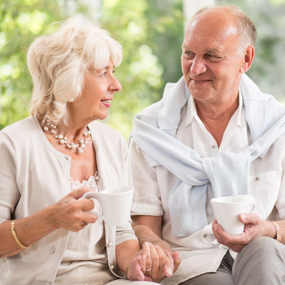 Elderly couple sitting outdoors, holding hands, drinking from white mugs, and smiling.