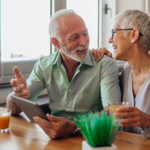 An older couple smiles, looking at a tablet together, arm around shoulder, drinks on the table.