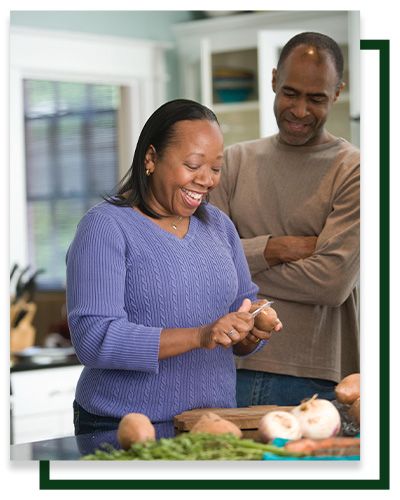 Woman peeling potatoes in a kitchen, smiling. Man watches, smiling, with arms crossed. Vegetables on counter.