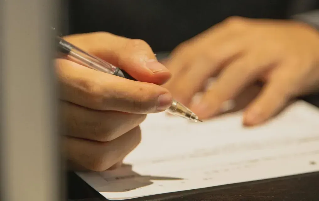 Person writing on a white paper with a clear pen at a dark desk.