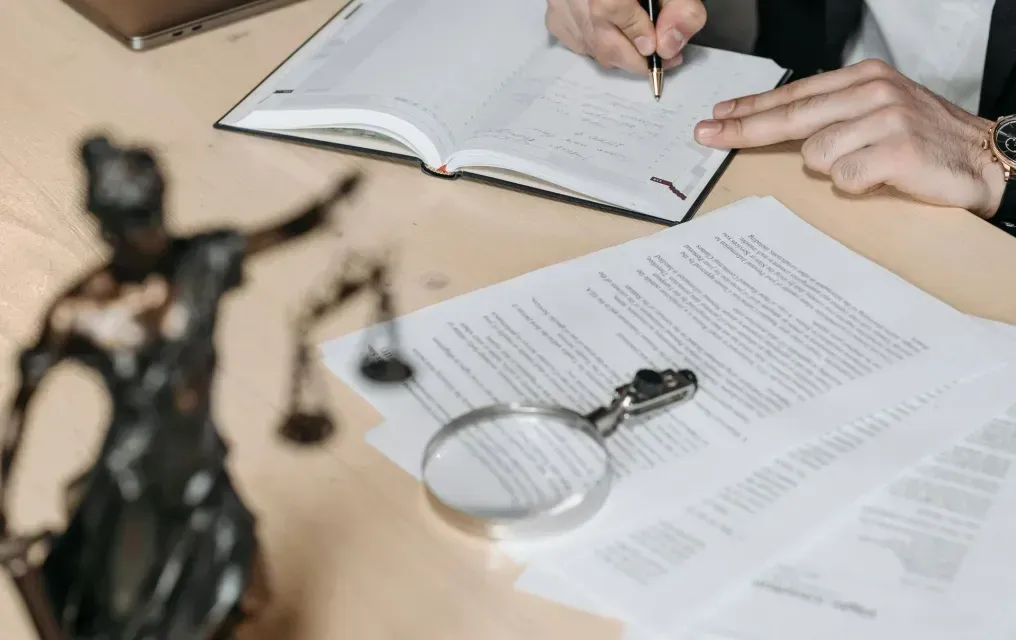 A person in a suit writes in a notebook at a desk with legal documents and a Lady Justice statue.
