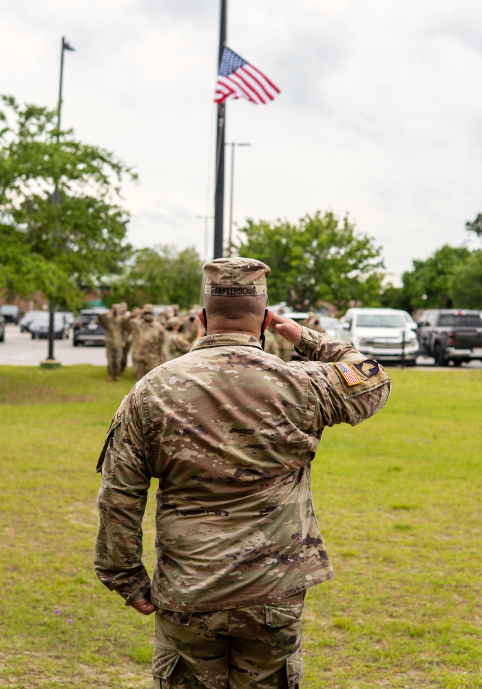 Soldier salutes American flag during a ceremony, other soldiers in the background, outdoor setting.