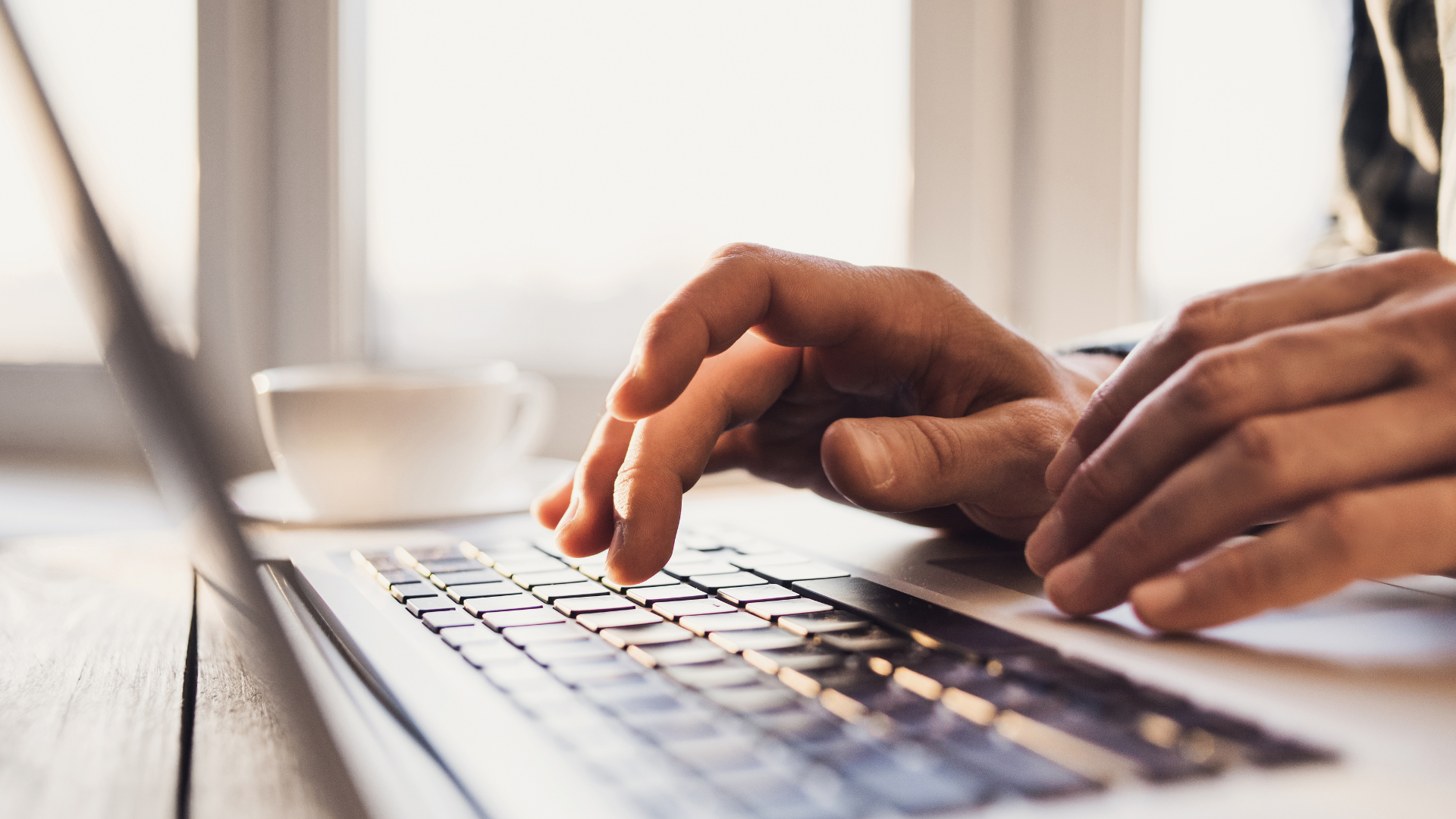 Hands typing on a laptop keyboard near a window with a coffee cup in view.