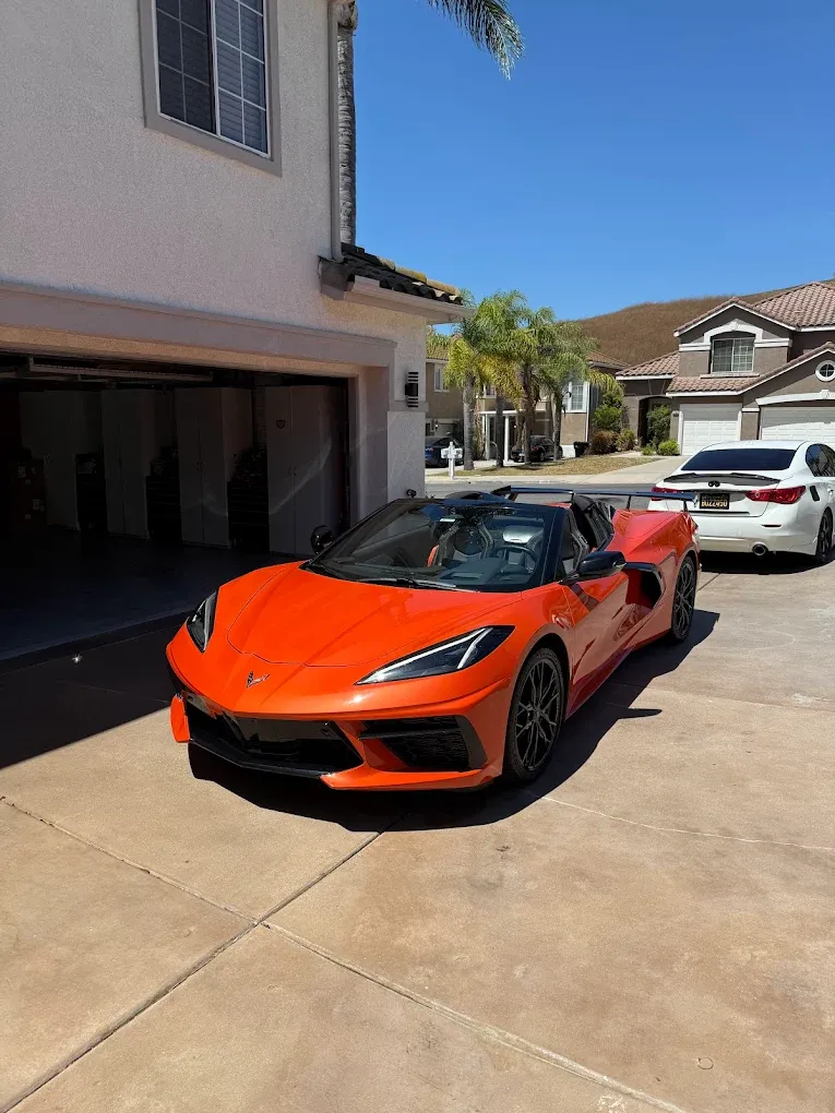 Orange sports car parked in a sunny driveway beside a house and white sedan
