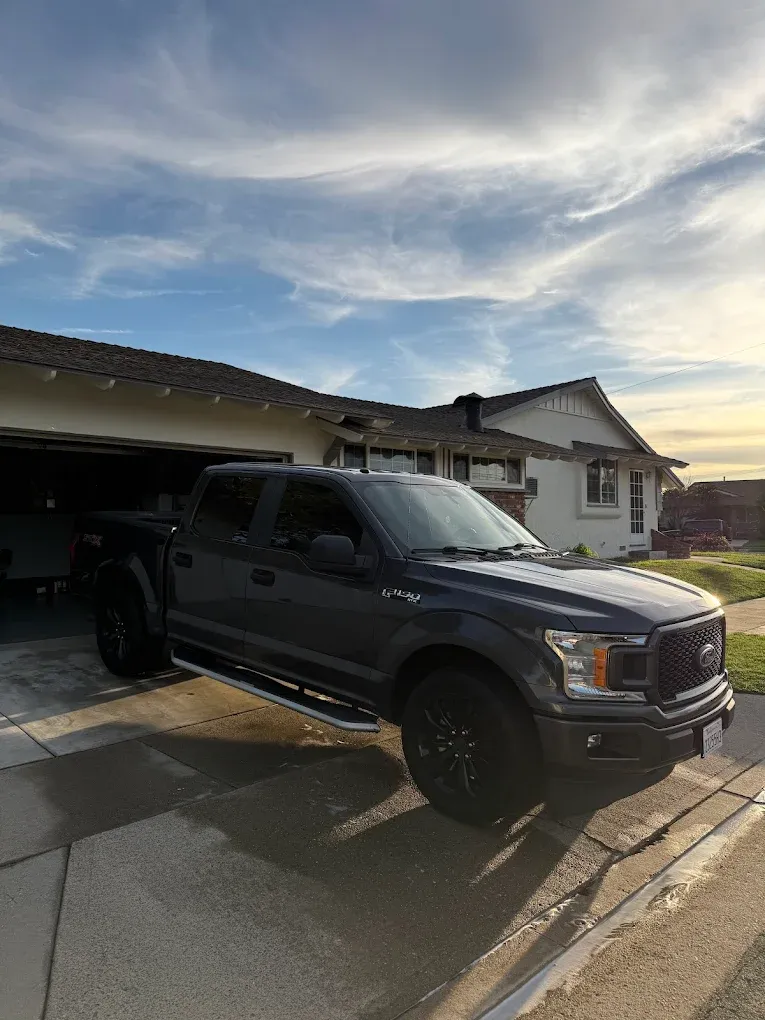 Black pickup truck parked in a driveway beside a suburban house at sunset.