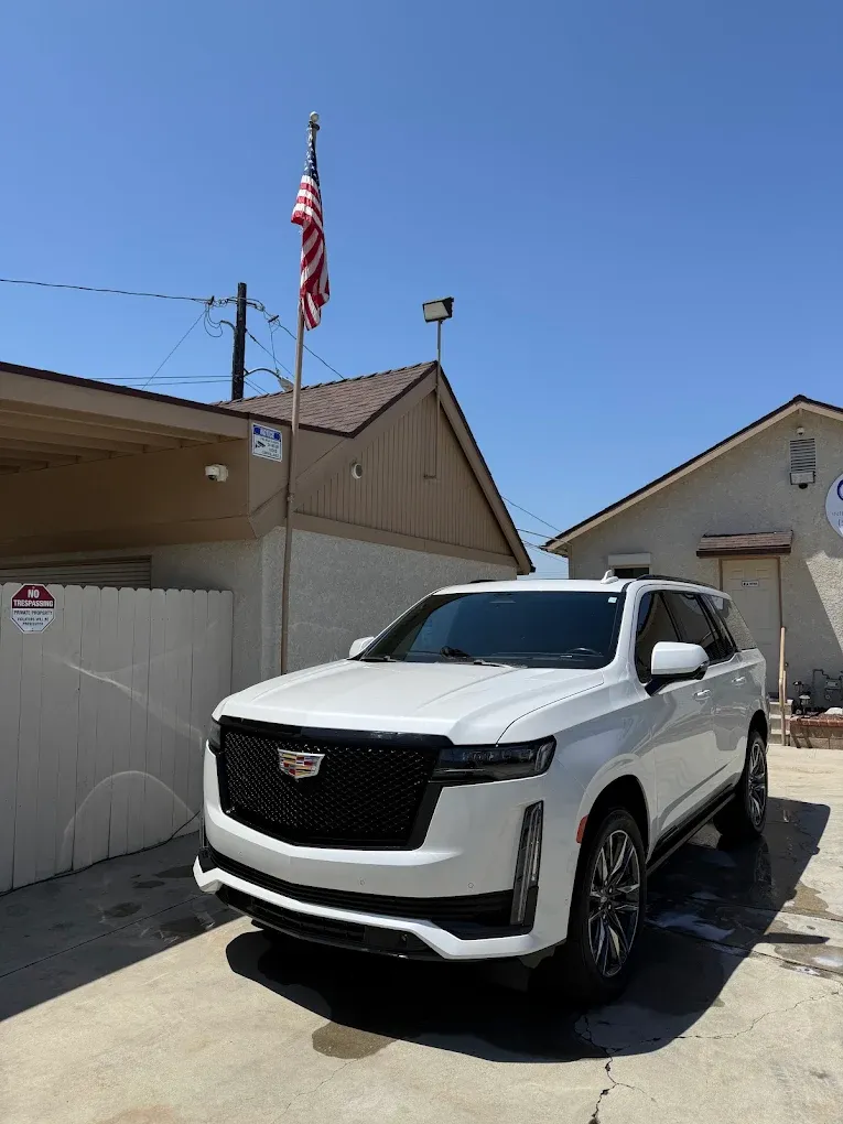 White Cadillac SUV parked in a sunny driveway beside a house with an American flag.
