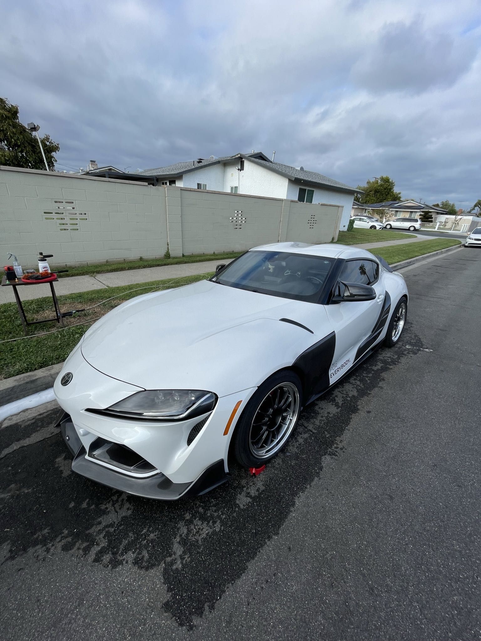 White sports car parked on a wet street beside a wall under cloudy skies