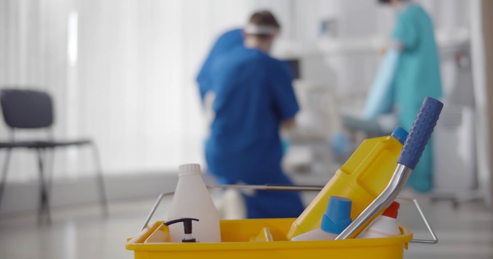 Cleaning supplies in foreground, medical staff working in background.